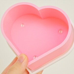 Puede incluir: Un molde de silicona rosa en forma de coraz&oacute;n con tres peque&ntilde;os agujeros en la parte inferior. El molde est&aacute; dise&ntilde;ado para hacer manualidades o hornear.