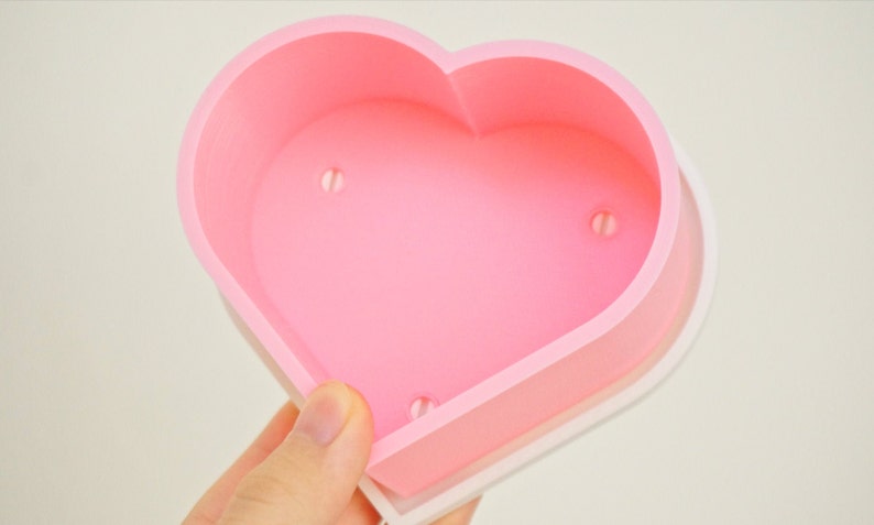 Puede incluir: Un molde de silicona rosa en forma de coraz&oacute;n con tres peque&ntilde;os agujeros en la parte inferior. El molde est&aacute; dise&ntilde;ado para hacer manualidades o hornear.