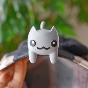 May include: A light grey cat-shaped accessory with black eyes and a black moustache is attached to a hat. The cat has a small tail and is made of a smooth material. The hat has a black and white pattern.