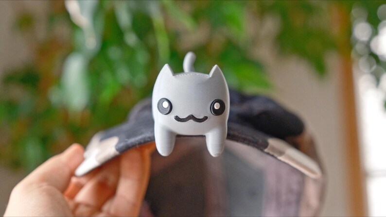 May include: A light grey cat-shaped accessory with black eyes and a black moustache is attached to a hat. The cat has a small tail and is made of a smooth material. The hat has a black and white pattern.