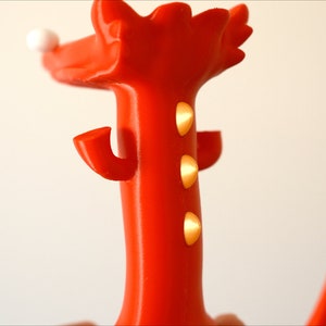 May include: A vibrant red decorative object with a whimsical design. It features a tall, slender body with three golden accents and curved arms. A white sphere sits atop the head, adding a playful touch. The object's bright color and unique shape make it a standout piece.