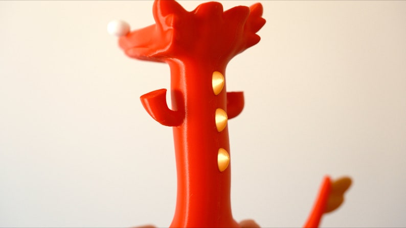May include: A vibrant red decorative object with a whimsical design. It features a tall, slender body with three golden accents and curved arms. A white sphere sits atop the head, adding a playful touch. The object's bright color and unique shape make it a standout piece.