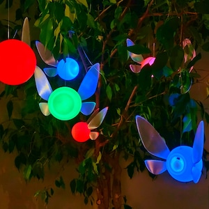 May include: Five hanging fairy lights shaped like the character Navi from the video game The Legend of Zelda. The lights are glowing in different colors: red, green, blue, and light blue.