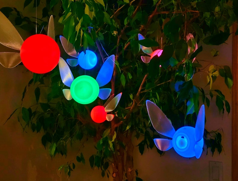May include: Five hanging fairy lights shaped like the character Navi from the video game The Legend of Zelda. The lights are glowing in different colors: red, green, blue, and light blue.