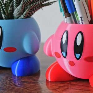 Puede incluir: Dos macetas de color rosa y azul impresas en 3D con forma del personaje de videojuego Kirby. La maceta azul tiene una peque&ntilde;a planta suculenta en su interior. La maceta rosa tiene una variedad de bol&iacute;grafos y l&aacute;pices en su interior.