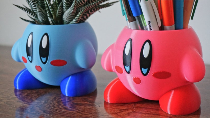 Puede incluir: Dos macetas de color rosa y azul impresas en 3D con forma del personaje de videojuego Kirby. La maceta azul tiene una peque&ntilde;a planta suculenta en su interior. La maceta rosa tiene una variedad de bol&iacute;grafos y l&aacute;pices en su interior.