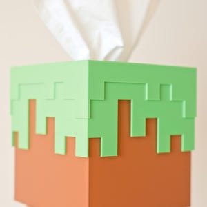 A cube tissue box cover inspired by the dirt grass block from the popular video game, Minecraft, is being held in front of a camera by a hand. It has bright green pixelated grass at the top half and the bottom half is brown.
