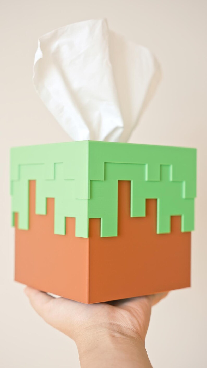 A cube tissue box cover inspired by the dirt grass block from the popular video game, Minecraft, is being held in front of a camera by a hand. It has bright green pixelated grass at the top half and the bottom half is brown.