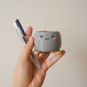 May include: A gray ceramic planter with a cute cartoon face and pink cheeks. The planter has four small, yellow legs and is being held by a hand.