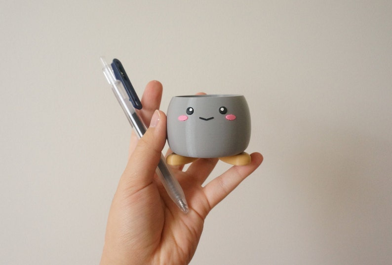 May include: A gray ceramic planter with a cute cartoon face and pink cheeks. The planter has four small, yellow legs and is being held by a hand.