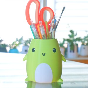 May include: A green frog-shaped pencil holder with a white belly and a happy face. The holder is filled with colourful pens, pencils, and paintbrushes.