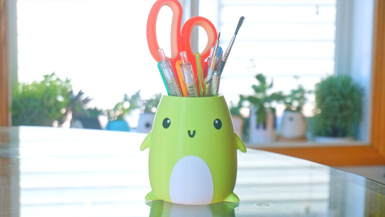 May include: A green frog-shaped pencil holder with a white belly and a happy face. The holder is filled with colourful pens, pencils, and paintbrushes.