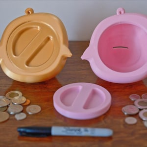 May include: Two piggy banks, one gold and one pink, with a coin slot on top. The gold piggy bank has a split design on the front. The pink piggy bank has a smooth front. A pile of coins is scattered on a wooden surface.