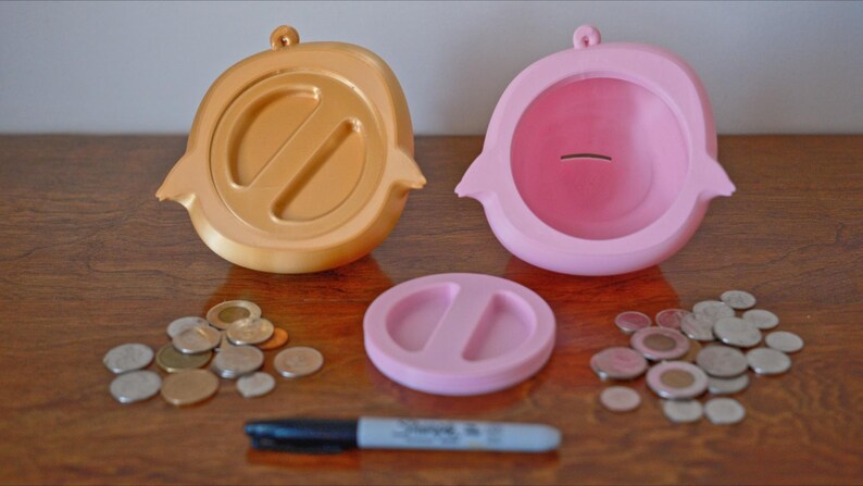 May include: Two piggy banks, one gold and one pink, with a coin slot on top. The gold piggy bank has a split design on the front. The pink piggy bank has a smooth front. A pile of coins is scattered on a wooden surface.