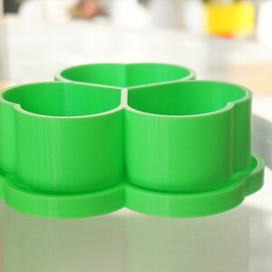May include: A bright green, three-sectioned container with a clover-shaped design. The container sits on a matching base, and the sections are open at the top. The object is made of plastic and is sitting on a glass surface.