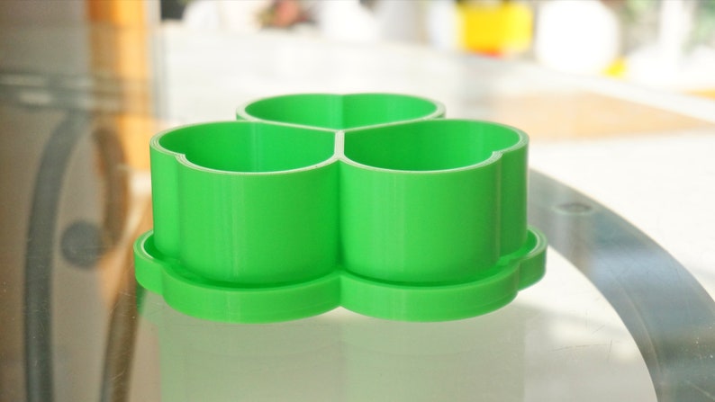 May include: A bright green, three-sectioned container with a clover-shaped design. The container sits on a matching base, and the sections are open at the top. The object is made of plastic and is sitting on a glass surface.