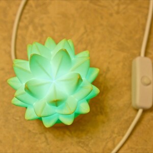 K&ouml;nnte beinhalten: Eine leuchtende, hellgr&uuml;ne Lotusbl&uuml;ten-Lampe mit wei&szlig;em Kabel und Ein-/Ausschalter. Die Lampe leuchtet und strahlt ein sanftes Leuchten aus. Der Hintergrund ist eine strukturierte, hellbraune Oberfl&auml;che.