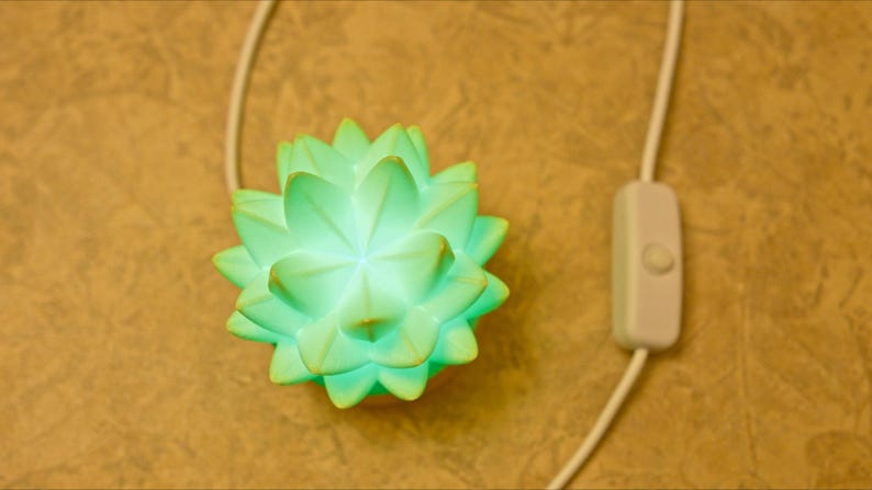 K&ouml;nnte beinhalten: Eine leuchtende, hellgr&uuml;ne Lotusbl&uuml;ten-Lampe mit wei&szlig;em Kabel und Ein-/Ausschalter. Die Lampe leuchtet und strahlt ein sanftes Leuchten aus. Der Hintergrund ist eine strukturierte, hellbraune Oberfl&auml;che.