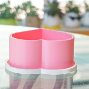 Puede incluir: Un macetero de cer&aacute;mica en forma de coraz&oacute;n rosa con un borde blanco. El macetero est&aacute; sobre una mesa de vidrio.