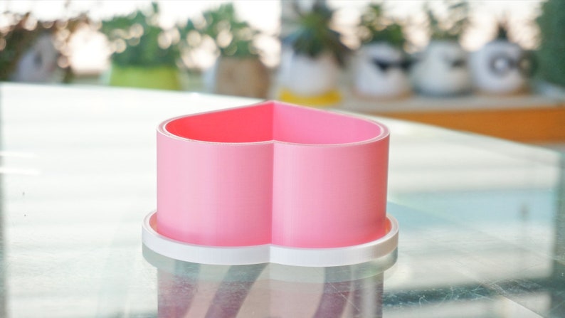 Puede incluir: Un macetero de cer&aacute;mica en forma de coraz&oacute;n rosa con un borde blanco. El macetero est&aacute; sobre una mesa de vidrio.