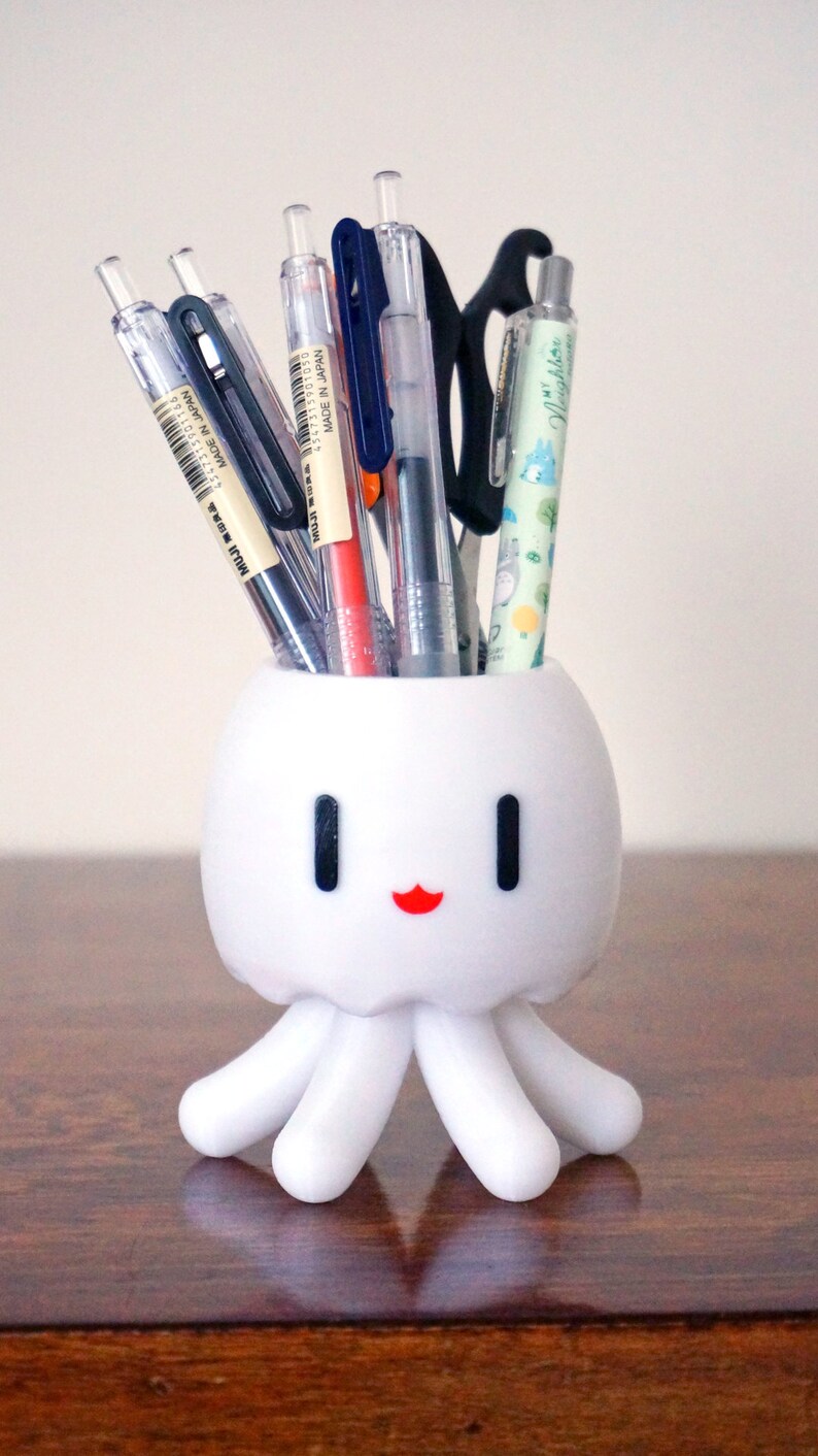 May include: White octopus-shaped desk organizer with a smiling face, holding a variety of pens and pencils, including a green pen with the text "Nightbloom" on it.
