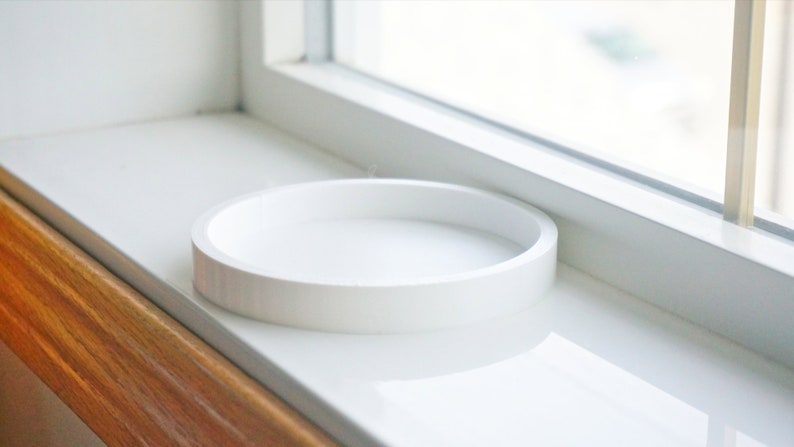 May include: A white ceramic round tray sits on a white windowsill.