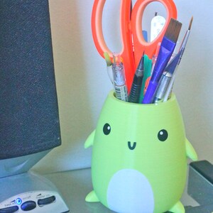 May include: A green frog-shaped pencil holder with a white belly and a happy face. The holder is filled with various writing utensils, including a pair of orange scissors, pens, and pencils.