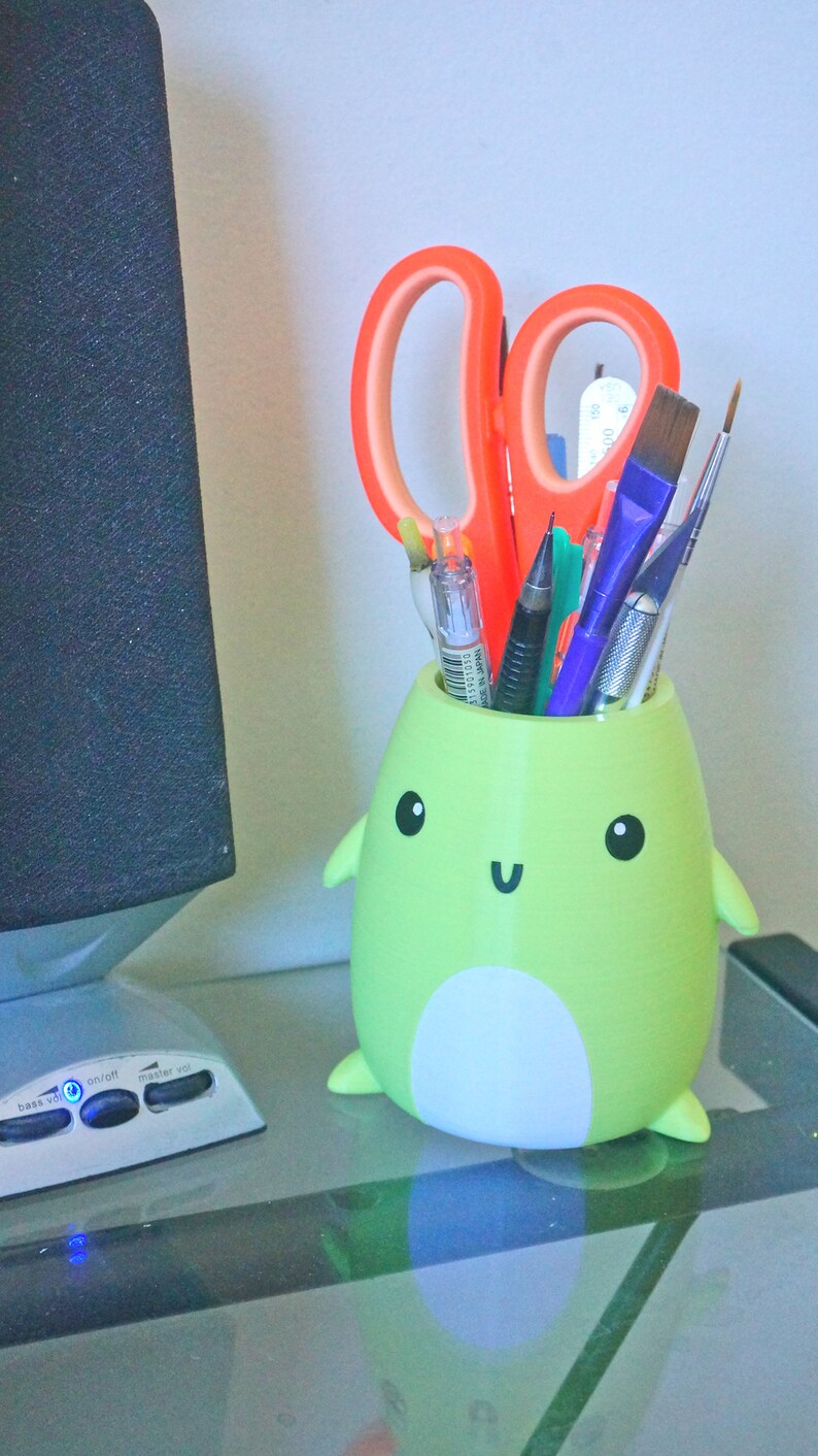 May include: A green frog-shaped pencil holder with a white belly and a happy face. The holder is filled with various writing utensils, including a pair of orange scissors, pens, and pencils.
