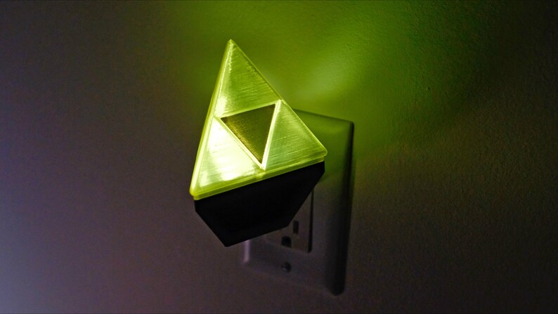May include: A yellow plastic night light shaped like the Triforce from the Legend of Zelda video game series. The night light is plugged into a standard electrical outlet.