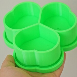 May include: A bright green, three-sectioned container in a clover shape. Each section is a heart shape. The container is held in a hand and appears to be 3D printed. The container is designed to hold small items.