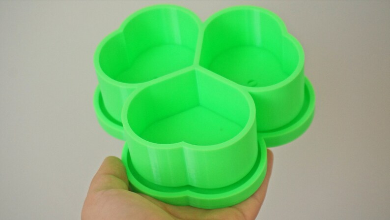 May include: A bright green, three-sectioned container in a clover shape. Each section is a heart shape. The container is held in a hand and appears to be 3D printed. The container is designed to hold small items.