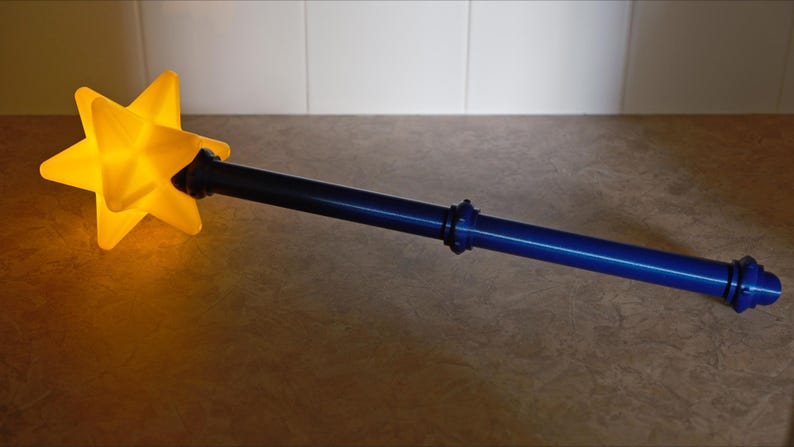 K&ouml;nnte beinhalten: Ein gelber sternf&ouml;rmiger Zauberstab mit blauem Griff. Der Stern leuchtet warm, der Griff ist dunkelblau. Der Zauberstab ist ein skurriles Objekt, perfekt f&uuml;r Kost&uuml;mspiele oder als dekoratives Element.