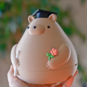 Puede incluir: Una figura en forma de oso beige con un gorro de graduaci&oacute;n y un diploma. El oso tiene una peque&ntilde;a flor rosa sujeta a su brazo. La figura est&aacute; sujeta en la mano de una persona.
