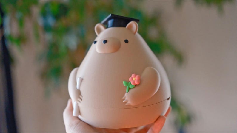 Puede incluir: Una figura en forma de oso beige con un gorro de graduaci&oacute;n y un diploma. El oso tiene una peque&ntilde;a flor rosa sujeta a su brazo. La figura est&aacute; sujeta en la mano de una persona.
