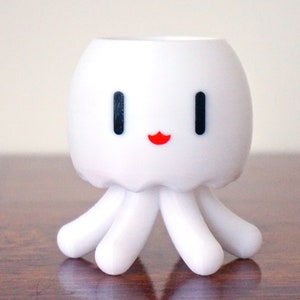 May include: White ceramic planter shaped like an octopus with a smiling face. The planter has black eyes and a red mouth.