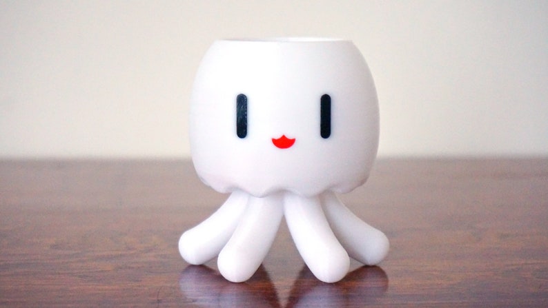 May include: White ceramic planter shaped like an octopus with a smiling face. The planter has black eyes and a red mouth.