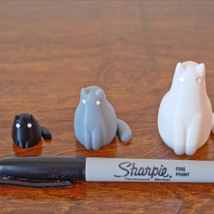 Może przedstawiać: Trzy ceramiczne figurki kot&oacute;w w r&oacute;żnych rozmiarach. Najmniejsza jest czarna, średnia jest szara, a największa jest biała.