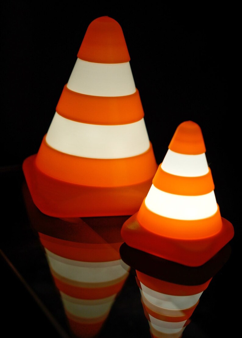 Op de afbeelding: Twee oranje en witte lampen in de vorm van verkeerskegels met een wit licht dat van binnenuit schijnt. De lampen staan op een zwart oppervlak.