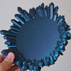 May include: A teal blue crystal-shaped dish with a round, flat interior. The dish is made of a textured material and has a unique, organic shape.