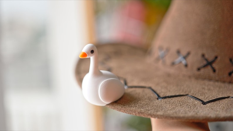May include: A white goose figurine with an orange beak sits on a brown cowboy hat. The hat has decorative stitching and a black brim. The figurine is a whimsical decorative item.
