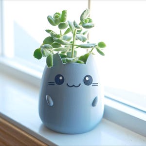 May include: A light blue cat-shaped planter with a smiling face and small arms and legs. The planter holds a small green succulent plant. The planter sits on a white windowsill.