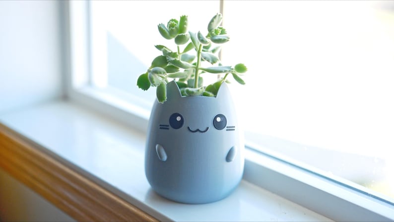 May include: A light blue cat-shaped planter with a smiling face and small arms and legs. The planter holds a small green succulent plant. The planter sits on a white windowsill.