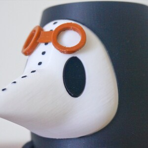 May include: A white and black figurine of a person wearing a black hooded robe, a white beak-like mask, and orange goggles. The figurine is made of plastic and has a textured surface.