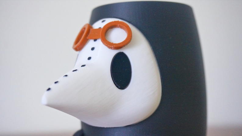 May include: A white and black figurine of a person wearing a black hooded robe, a white beak-like mask, and orange goggles. The figurine is made of plastic and has a textured surface.