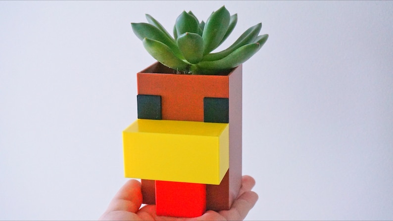 K&ouml;nnte beinhalten: Ein brauner, gelber und roter Pflanzer in Form eines Minecraft-Huhnkopfes mit einer gr&uuml;nen Sukkulente, die darin w&auml;chst.
