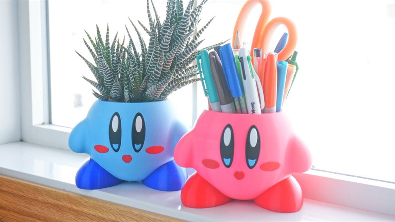 Puede incluir: Dos macetas impresas en 3D de color rosa y azul con forma del personaje Kirby de la serie de videojuegos Kirby. La maceta azul tiene una peque&ntilde;a planta suculenta en su interior. La maceta rosa est&aacute; llena de bol&iacute;grafos y l&aacute;pices.