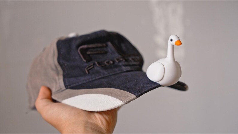May include: A baseball cap with a white brim, gray and navy blue panels, and the word "FLORIDA" embroidered on the front. A small white goose figurine with an orange beak is perched on the cap's brim.