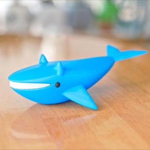 May include: A bright blue whale figurine with a white mouth and black eye details. The whale has fins and a tail, and is sitting on a wooden surface. The figurine is a decorative item.