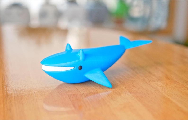 May include: A bright blue whale figurine with a white mouth and black eye details. The whale has fins and a tail, and is sitting on a wooden surface. The figurine is a decorative item.