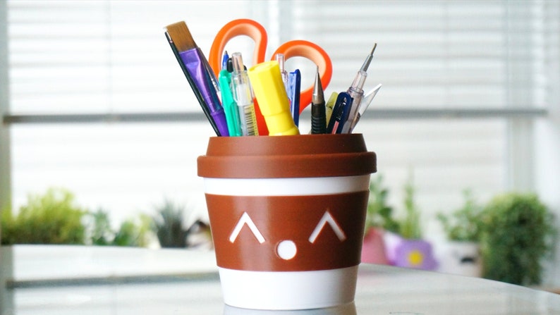 May include: A brown and white ceramic coffee cup with a cute face design, used as a pencil holder. It is filled with various writing utensils, including pens, pencils, and a paintbrush.